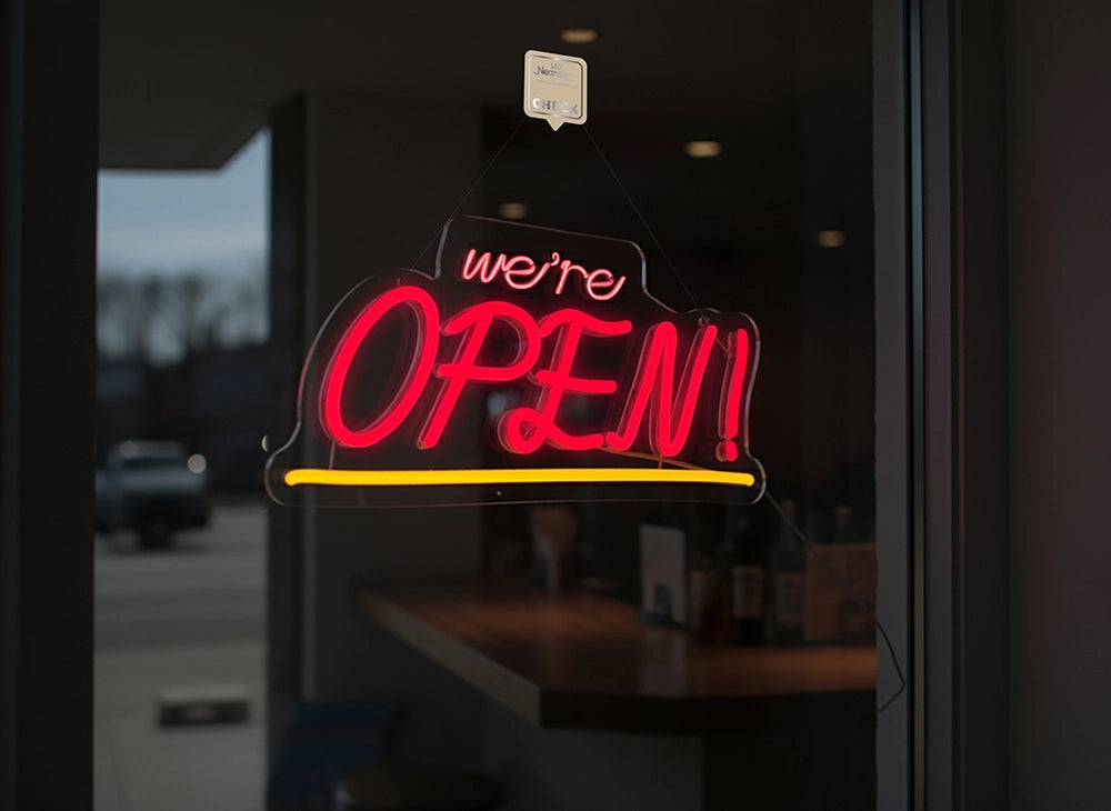 Neon 'we're OPEN!' sign on a glass door with a blurred street view in the background.